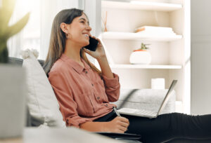 Laptop, phone call and writing with freelance woman on sofa in living room of home for communication. Computer, smile and thinking with happy person talking on mobile in apartment for remote work.